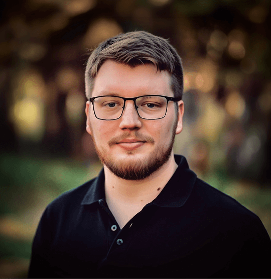 A man with glasses and a beard wears a black polo shirt, standing outdoors with a blurred green and brown background. He appears calm and relaxed.